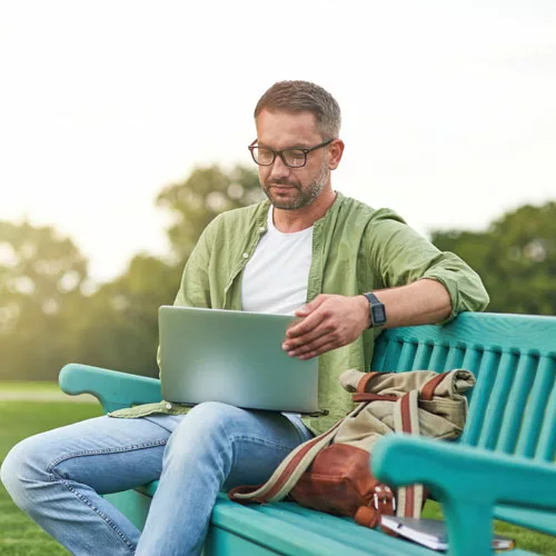 A man with glasses, comfortably seated on a park bench, engrossed in viewing content on his laptop, capturing the blend of nature and technology, and his focused demeanor in an outdoor setting.
