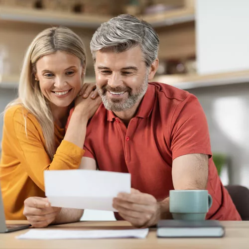 A man and woman, both wearing pleasant smiles, closely examining and discussing paperwork together, reflecting their shared interest, positive interaction, and the significance of the information they're reviewing.