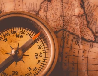 An aerial close-up view of a compass, its needle pointing to a direction, placed overlapping a detailed map, emphasizing the tools of navigation, exploration, and the intricate geography depicted on the map.