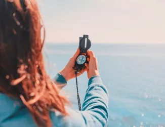 A woman, with her hands outstretched, holding and intently looking at a compass, set against the backdrop of a vast water body, reflecting her sense of exploration, direction-seeking, and the vastness of the natural setting.
