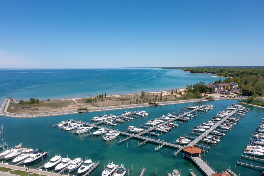 A scenic landscape view of a Michigan harbor, showcasing boats docked peacefully, with the shimmering blue-green waters reflecting the sky and surrounding area, capturing the serenity and beauty of the lakeside setting.