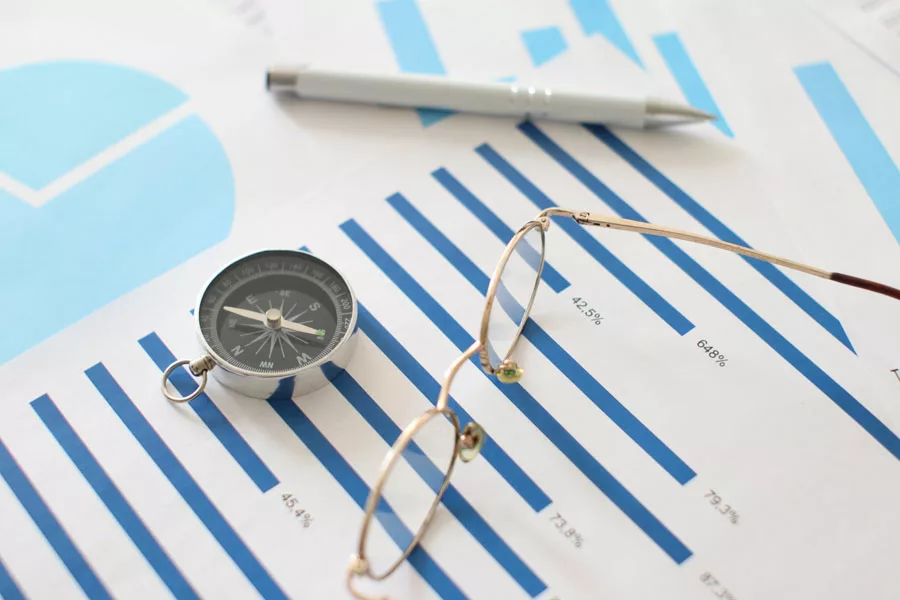 An aerial view of a bar graph on paperwork, complemented by a compass, pen, and a pair of glasses laid on top, emphasizing the analytical and navigational nature of the task and the tools aiding the process.